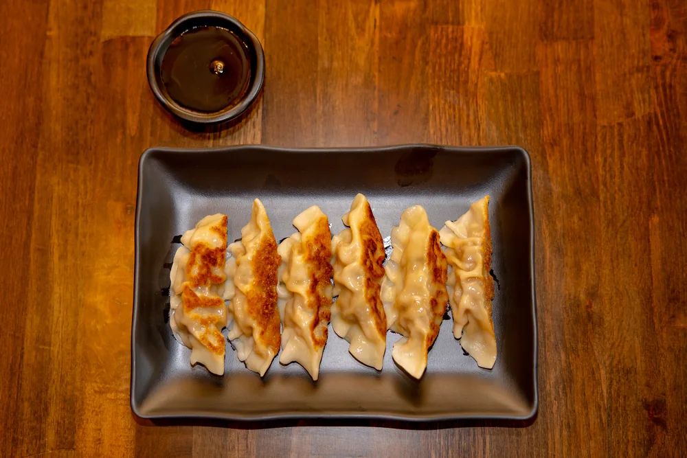 Six pan-fried gyoza in a row on a black plate with a bowl of dipping sauce