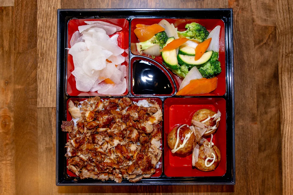 Bento box with chopped glazed chashu over rice, takoyaki, vegetables and pickles