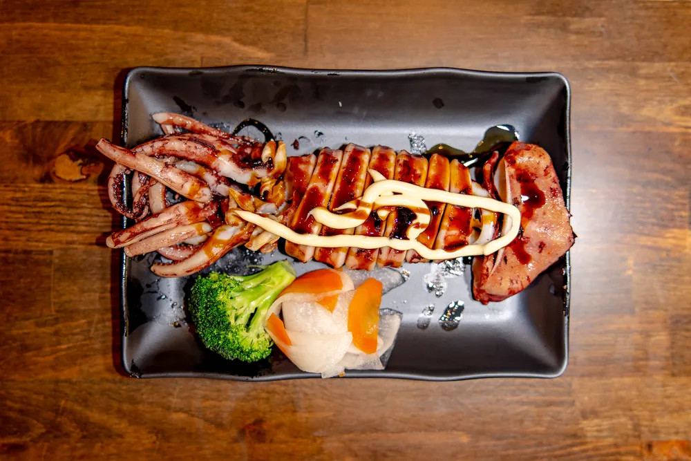 A grilled whole squid sliced and glazed, with broccoli and pickled daikon on a black plate
