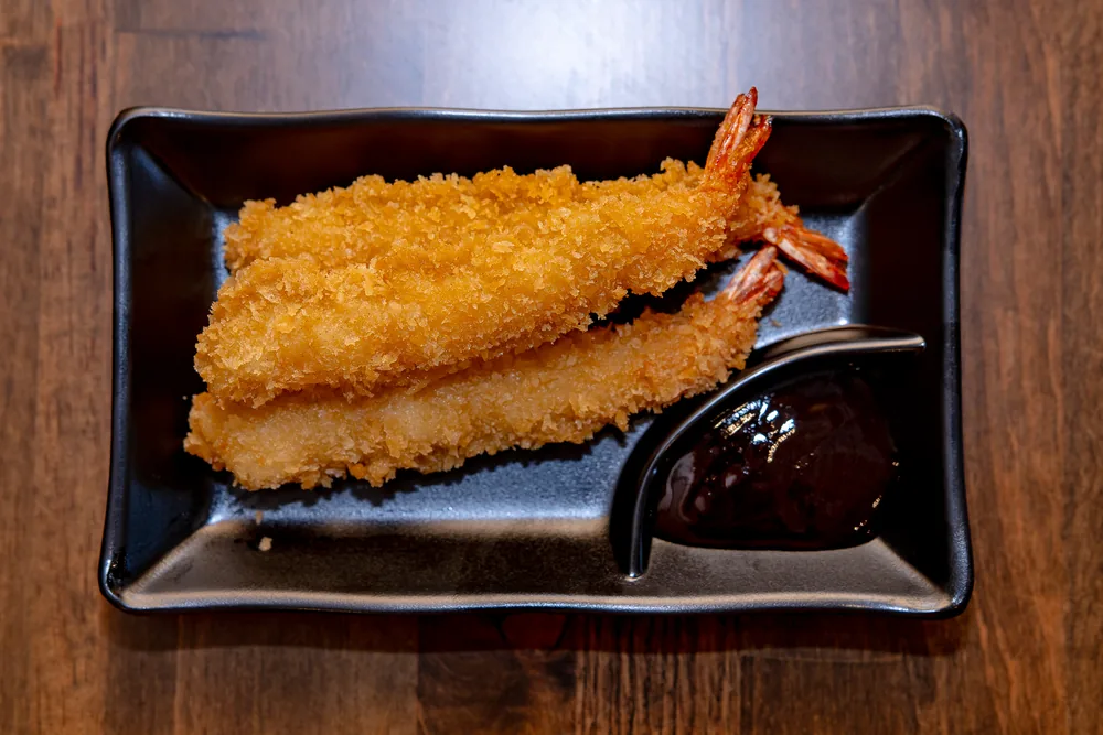 Panko-fried shrimp on a black plate with a dish of dark dipping sauce