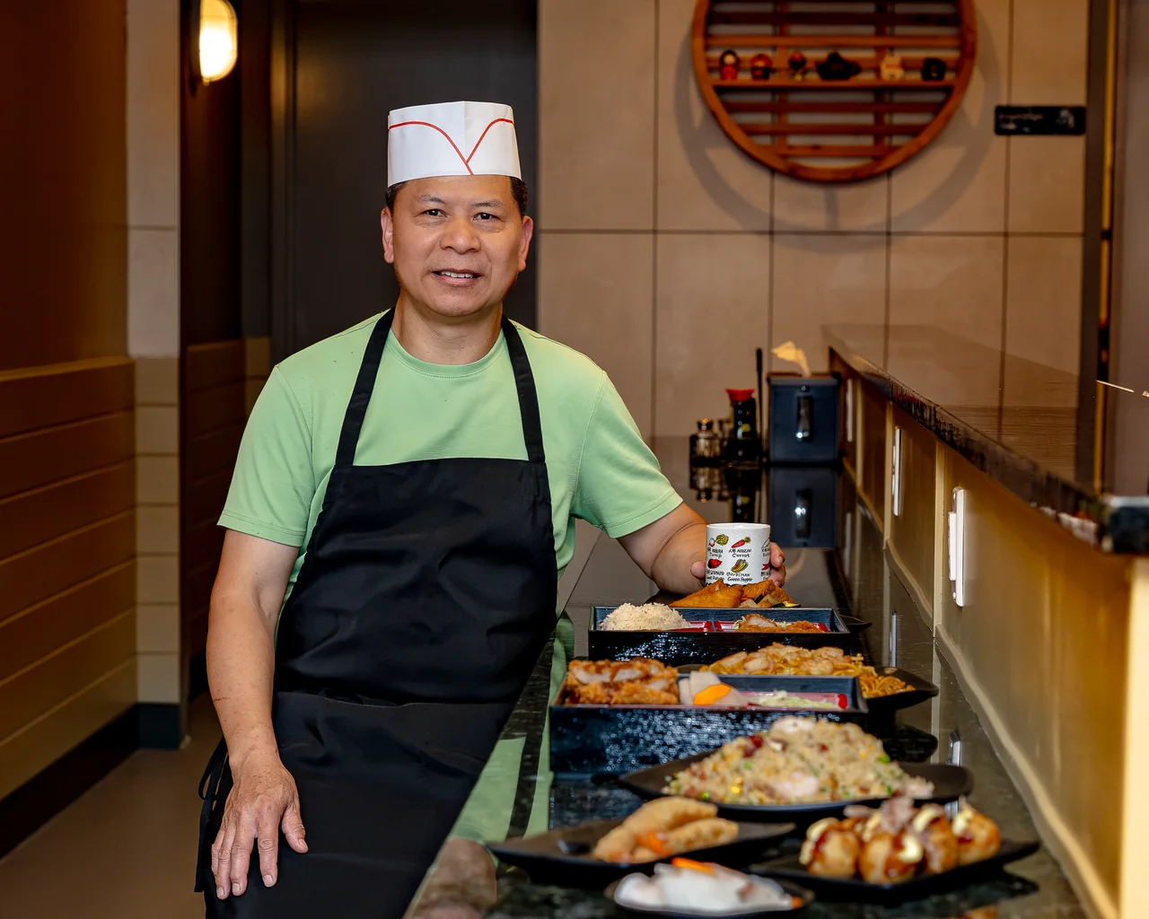A chef at Momiji in an apron and paper hat standing at the counter beside a row of freshly plated bento boxes, fried rice, yakisoba and small plates