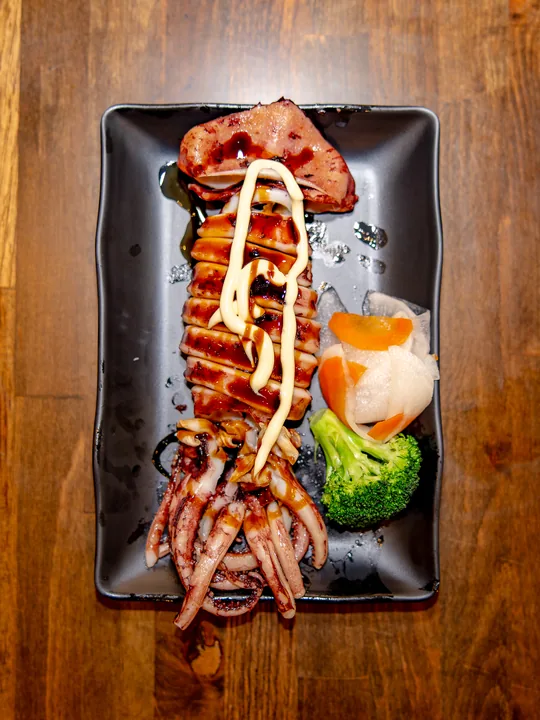 A grilled whole squid on a black plate, glazed and drizzled with Japanese mayo, served with broccoli and pickled daikon
