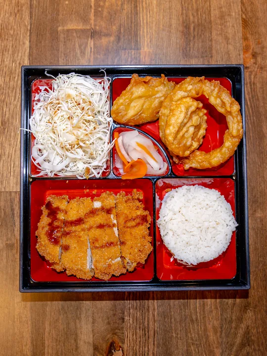 A bento box with a sliced breaded chicken katsu cutlet, white rice, shredded cabbage, pickled daikon and fried side pieces