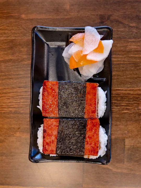Two pieces of grilled spam musubi wrapped in nori on a black plate with pickled daikon and carrot