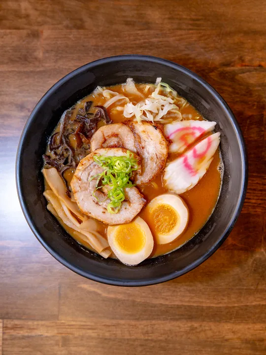 A bowl of ramen in a black bowl with two slices of chashu pork, a halved soft-boiled egg, fish cake, bamboo shoots, wood-ear mushroom and cabbage